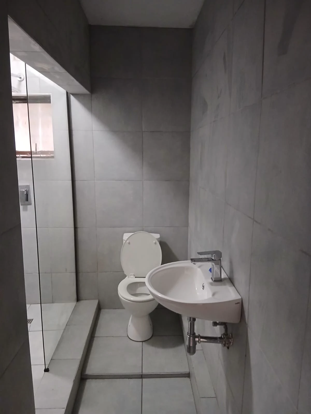 A finished bathroom tiled floor to ceiling in large grey tiles, with a toilet, basin and glass shower screen