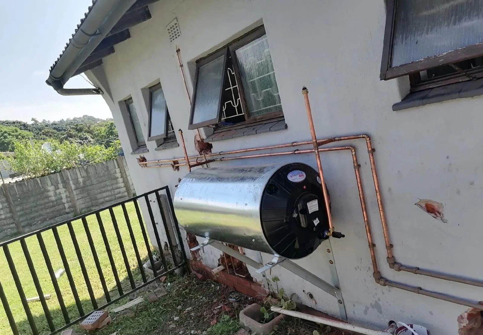 A geyser mounted on an exterior wall with a full copper pipe run above and around it