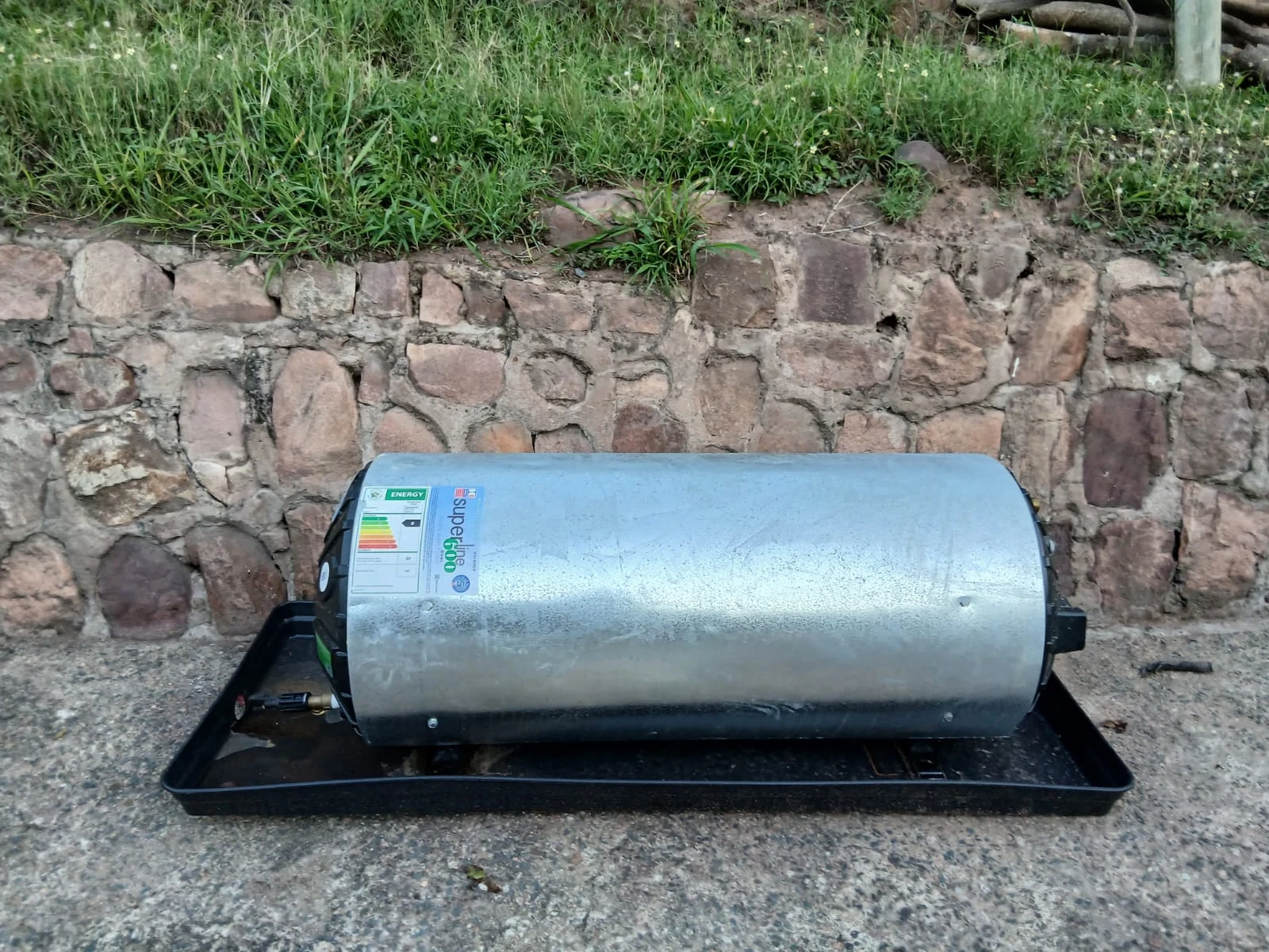 A new galvanised geyser standing on a black drip tray against a stone wall