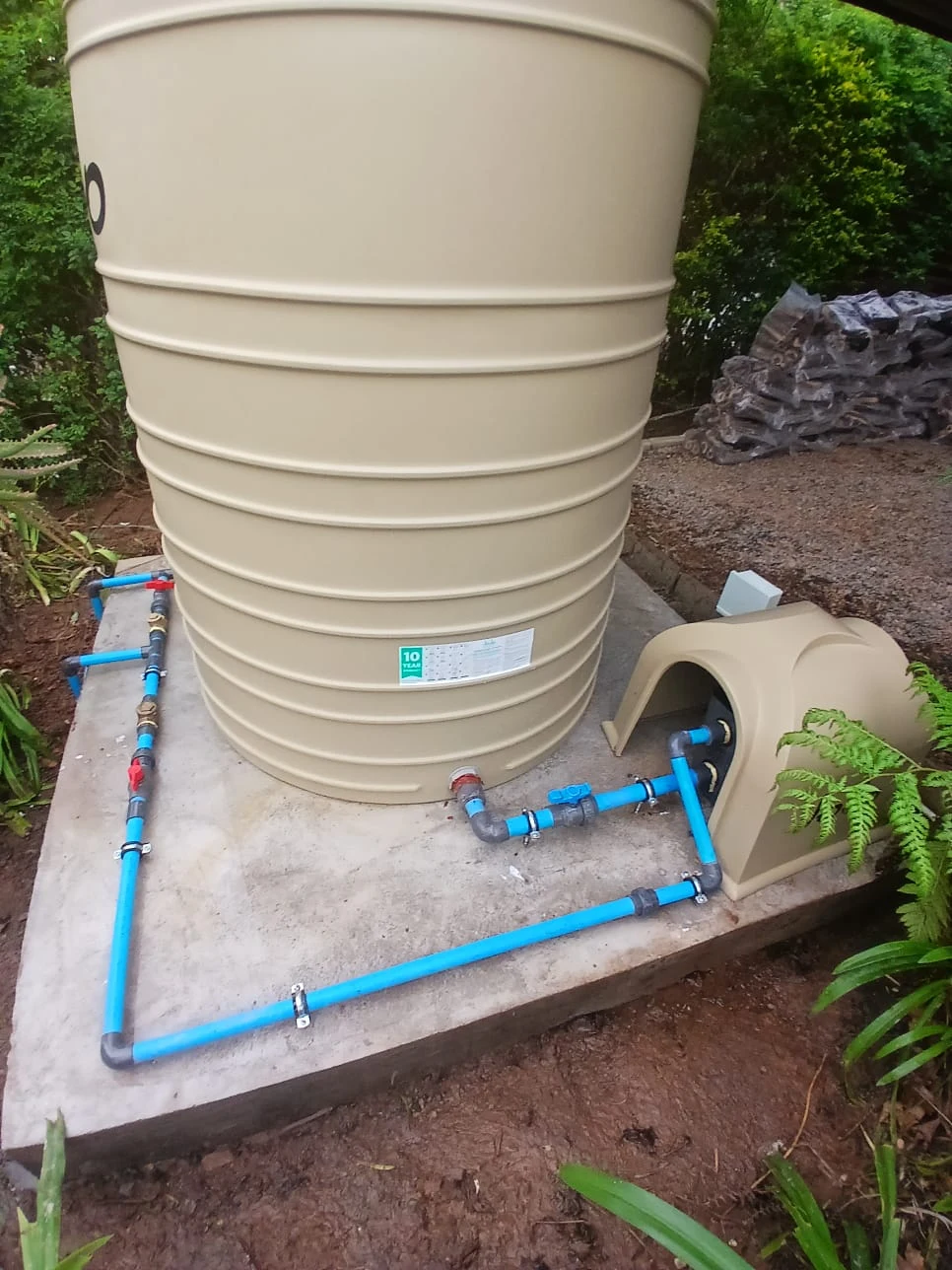 A large water storage tank on a concrete base with blue pipework and valves running across it, and a moulded cover housing the pump beside it