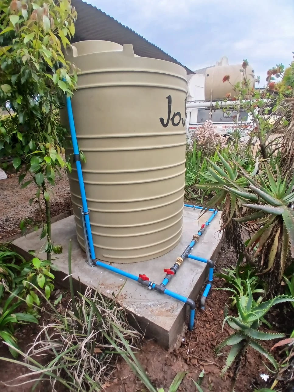 A water storage tank newly set on a concrete base with blue pipework and isolating valves, a delivery truck carrying another tank behind it